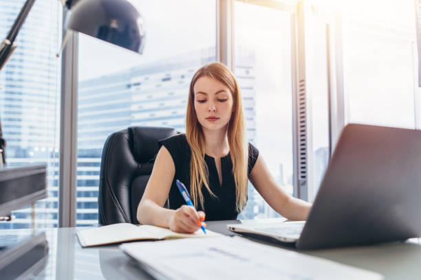 istock Female chief executive sitting at her desk taking notes in datebook writing with pen and using her computer in modern office building 891318232
