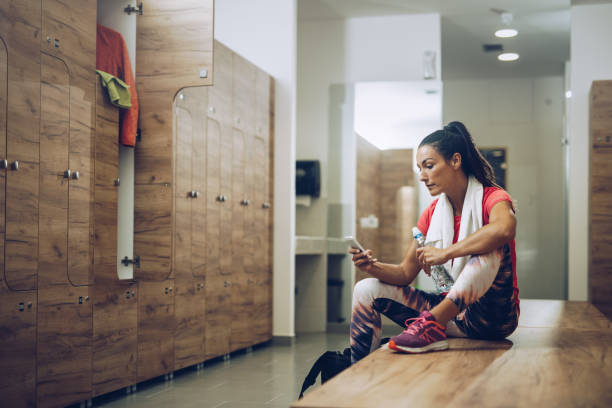Young athletic woman sitting on a bench in dressing room and using smart phone.