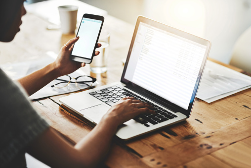 Closeup shot of an unidentifiable businesswoman using a cellphone and laptop while working in an office