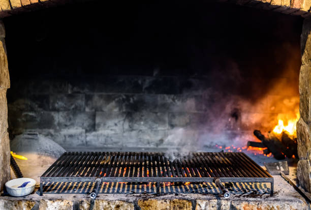 Professional big stone paved barbecue fireplace grill in a restaurant. Grilling on charcoal or coal with big grill grates or grids, and burning wood in the background.