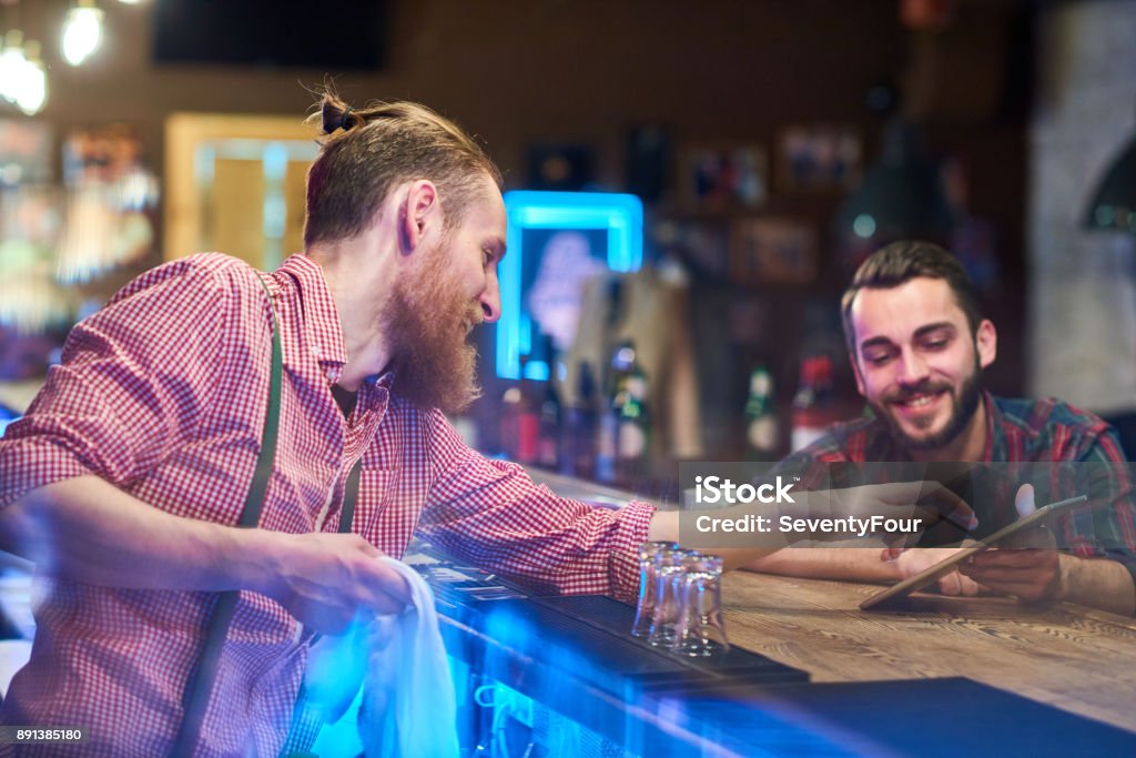 Bartender Helping Guest Choosing Drinks Side view portrait of modern bearded bartender talking to client at bar counter in pub, helping him choose drinks via digital tablet Bartender Stock Photo