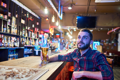 Portrait of modern bearded man in sitting at bar counter in pub watching sports match on TV  and drinking beer, copy space