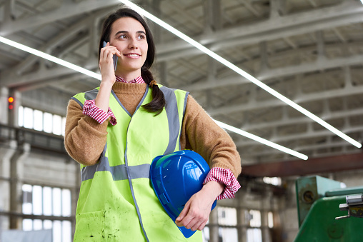 Smiling young technician wearing reflective jacket looking away while talking to her colleague on smartphone, interior of spacious production department on background