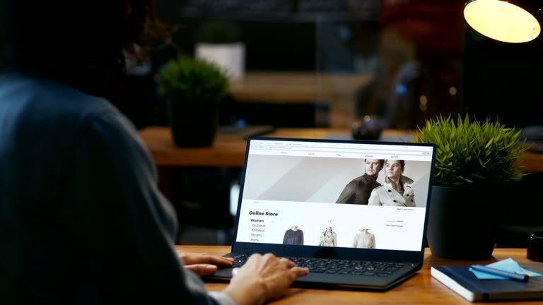 Woman Sitting at Her Desk with the Laptop Looks at Online Retail Clothing Store Website. Site Shows Various Male and Female Stylish Clothes, New Collection.