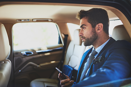 Shot of a focused young businessman using his cellphone and contemplating while being seated in the back of a car