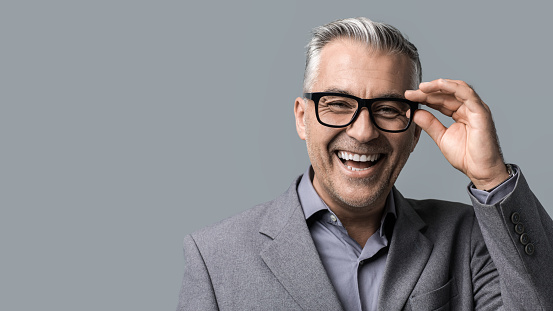 Smart mature businessman with glasses posing on gray background, he is smiling at camera