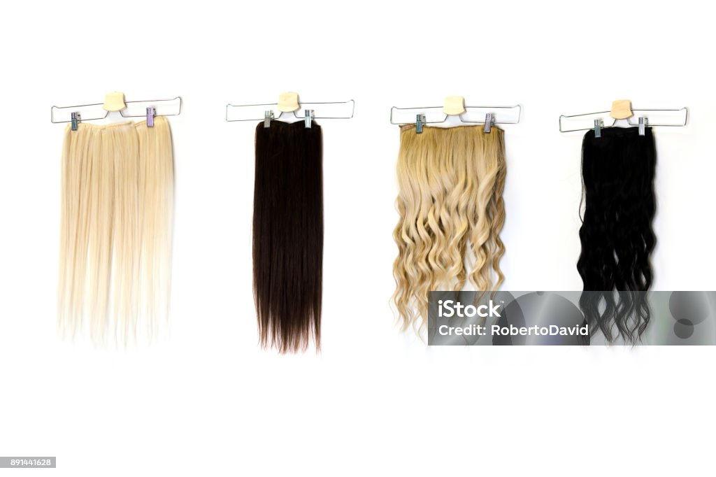 Colored hair extensions on clips in salon Natural human hair extensions hung on a white wall in the beauty salon, space for text. Multicolor straight and curly tape in colorful remy human hair extensions. Wig Stock Photo