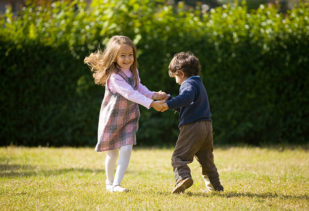 Back view of Asian girl child and boy kid wearing cap hat with hand in hands and waving palm hello or bye while standing in the garden.