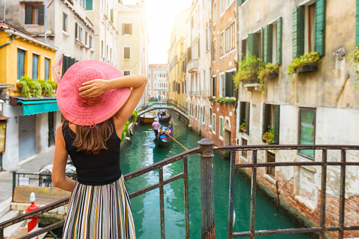 Elegant woman with red sunhat enjoys the view to a canal with passing by gondola in Venice, Italy