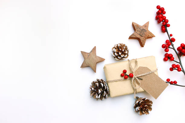 Christmas festive styled stock image. Floral frame composition with gift box, craft paper tag, pine cones, eucalyptus branches, wooden stars and holly berries on white wooden background. Flat lay, top view with empty copy space