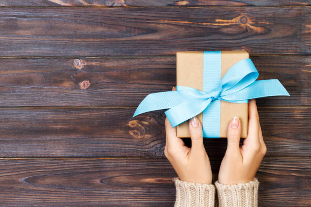 woman hands give wrapped valentine or other holiday handmade present in paper with blue ribbon. present box, decoration of gift on white wooden table, top view with copy space - hand wrap imagens e fotografias de stock