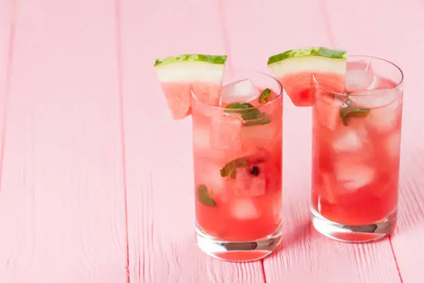 Watermelon lemonade with ice and mint leaves. Homemade lemonade of ripe berry with red and green ripes. Glass of cold watermelon tea. Refreshing summer drink. Cocktail on a wooden background.