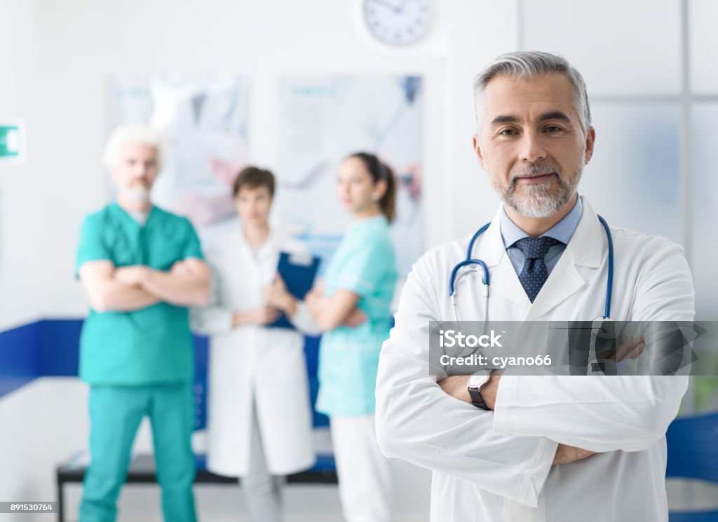 Confident doctor posing at the hospital Confident smiling doctor posing and the hospital with arms crossed and medical team working on the background Doctor Stock Photo