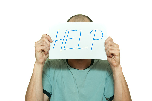 Young depressed man suffering from anxiety and feeling miserable holding help sign on paper in his hands and leaning on the white wall partially isolated