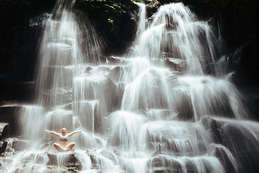 Travel in Bali jungle. Beautiful young woman sitting in yoga pose on rock under falling spring water, enjoy tropic cascade waterfall. Nature day trip, walking adventure, fun on family summer vacation