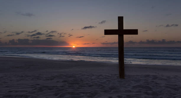 An old cross on a sand dune next to the ocean with a calm sunrise stock photo
