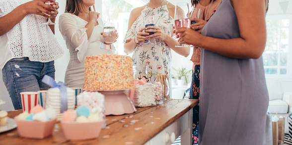 Cropped shot of cake and sweet food on table with women standing around holding drinks at baby shower party. Female friends enjoying baby shower party