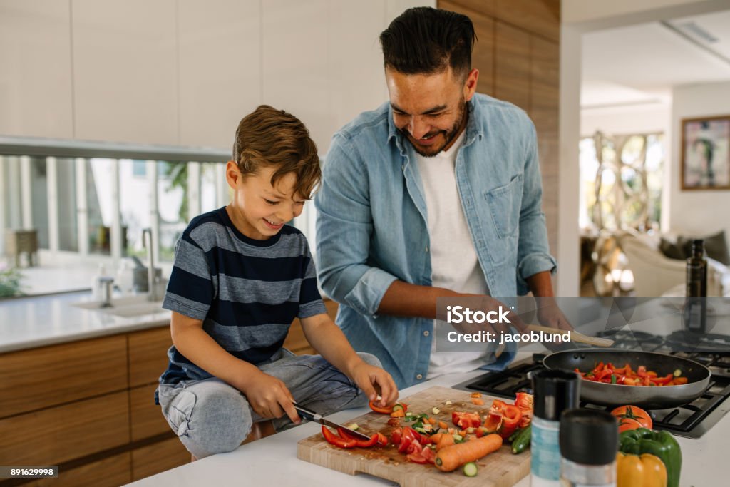 Father and son preparing food in kitchen Little boy cutting vegetables while his father cooking food in kitchen. Father and son preparing food at home kitchen. Cooking Stock Photo