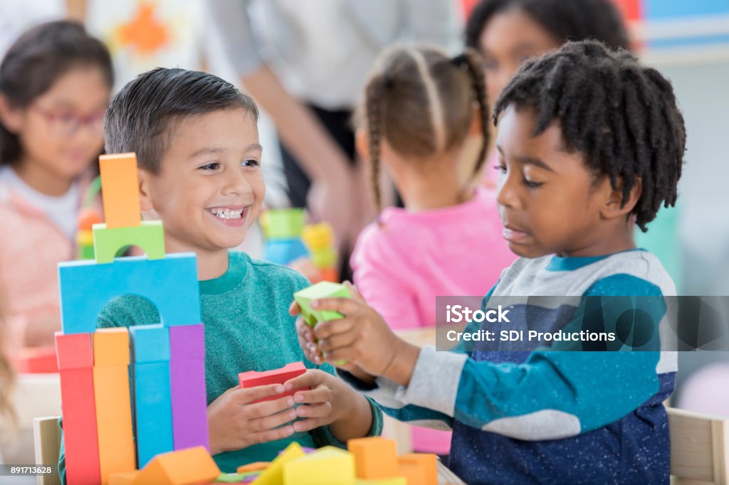 Kindergarten students play with toy blocks Diverse kindergarten schoolboys enjoy building towers with foam blocks. They enjoy playing with one another. Students are playing in the background. Sharing Stock Photo