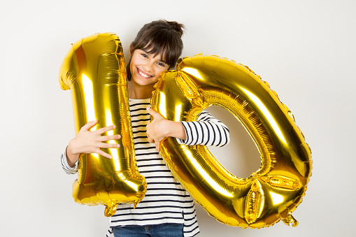 Happy little girl holding two golden balloons making the 10 number on white background. 10th anniversary celebration party.