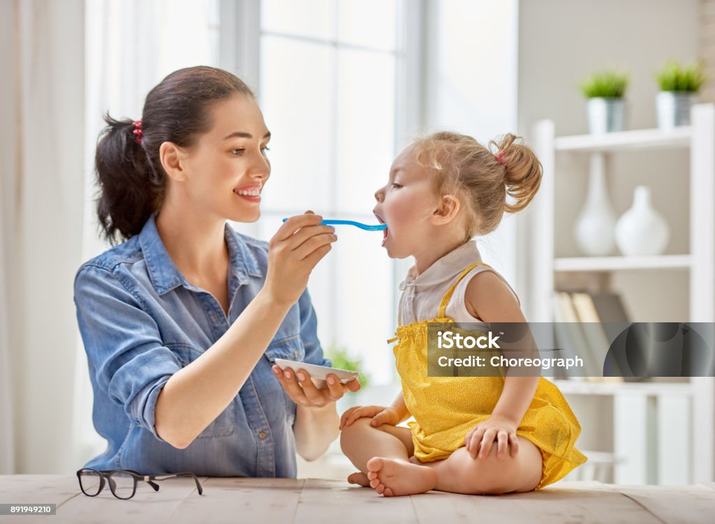 mother feeding her child Happy young mother feeding her baby girl with a spoon at home. Baby - Human Age Stock Photo