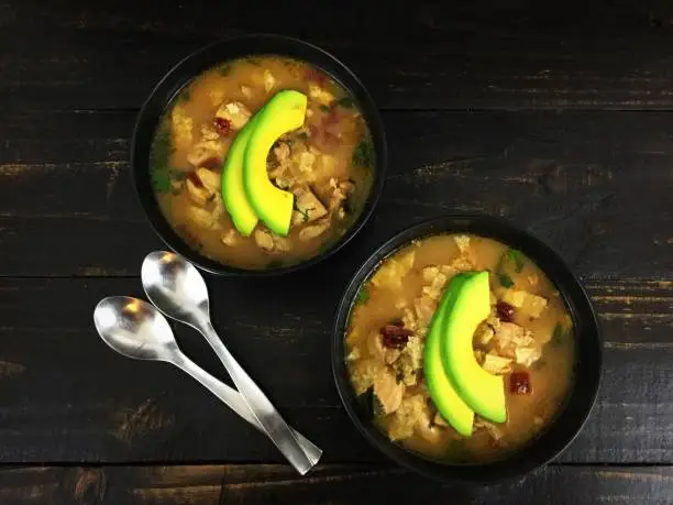 Two bowls of chicken soup topped with sliced avocado and shown with spoons