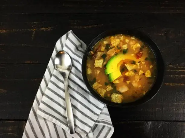 A bowl of chicken soup topped with sliced avocado and shown with a spoon and napkin