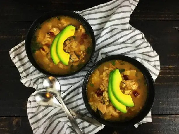 Two bowls of chicken soup topped with sliced avocado and shown with spoons