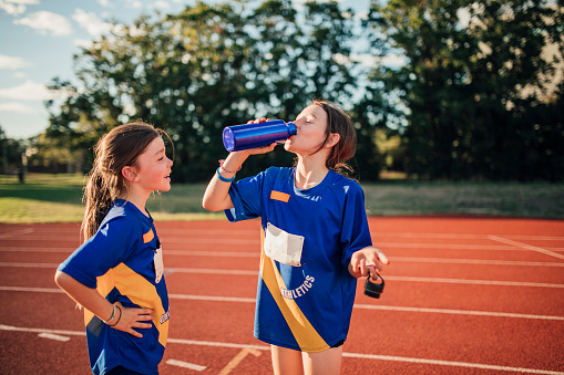 Two little girls are having a water break on the running track at athletics club. One girl is standing with her hands on hips, the other is taking a big gulp of water from  bottle.