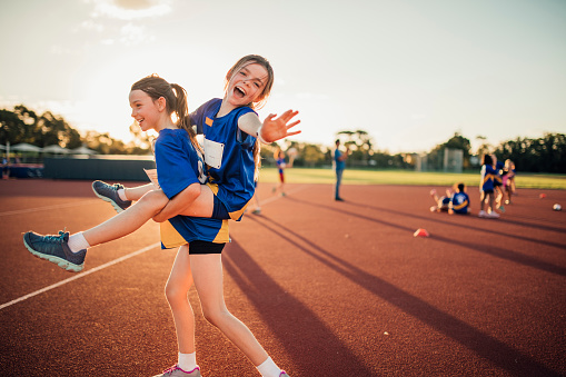 Two girls are messing around at athletics club on the running track. One is giving the other a piggy back and she is reaching out for the camera.