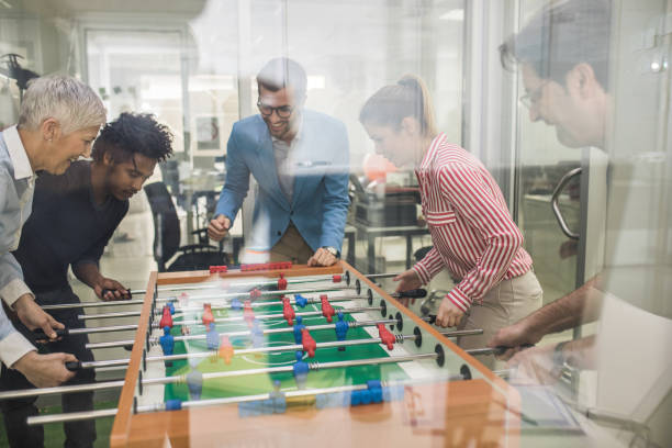 Playful business people having fun while playing foosball on a break in the office. One of them is cheering. The view is through glass.