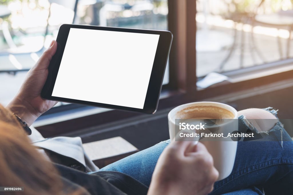 Mockup image of woman's hands holding black tablet pc with white blank screen and coffee cup of hot latte sitting in modern cafe Digital Tablet Stock Photo