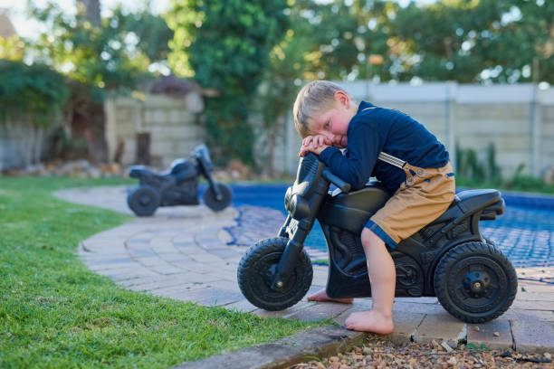 Toddler holding a black balance bike handlebar, surrounded by green plants and colorful flowers in a garden setting.