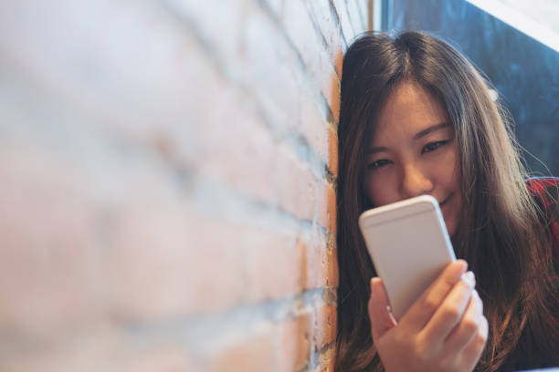 istock Closeup image of a smiley beautiful Asian woman leaning on brick wall while using and looking at smart phone in modern cafe 891996644