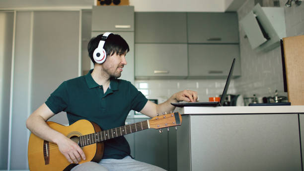 istock Cheerful young man with headphones sitting at kitchen learning to play guitar using laptop computer at home 892092430