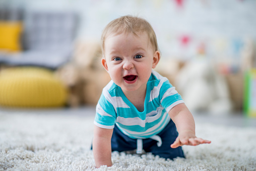 A Caucasian baby boy is crawling towards the camera while laughing. He is giggling and looking at the camera. He is at a daycare center.