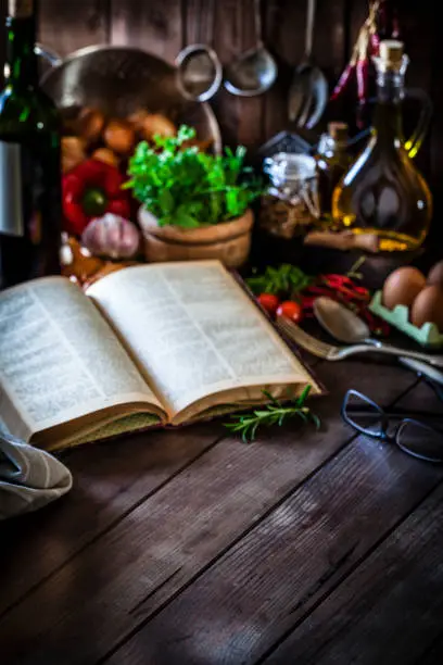 Vertical shot of an old open cookbook on rustic kitchen table. The cookbook is at the center of the frame leaving useful copy space for text and/or logo or for placing any product or food. At background are visible out of focus some herbs, spices, olive oil bottles and some kitchen utensils. Low key DSRL studio photo taken with Canon EOS 5D Mk II and Canon EF 100mm f/2.8L Macro IS USM