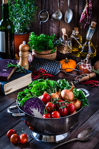 Vertical shot of fresh organic vegetables on rustic kitchen table. The composition includes lettuce, bell peppers, tomatoes, edible mushrooms, cauliflower, cucumber, broccoli, onion, carrot, corn, and chili peppers. The vegetables are at foreground in a colander. An old cookbook is partially visible at the left. Low key DSRL studio photo taken with Canon EOS 5D Mk II and Canon EF 100mm f/2.8L Macro IS USM