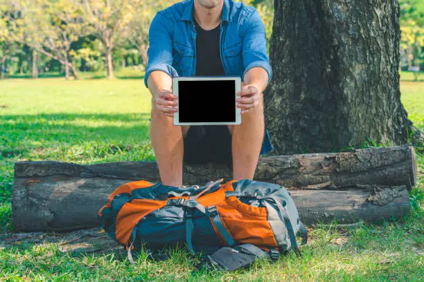 Man holding a digital tablet sitting on a log in park. A backpack on the grass