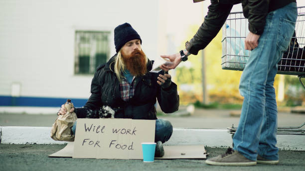若い男は、ホームレスの人、通りで彼の乞食お酒と買い物カゴの近くに座って、いくつかの食糧を与えること - homelessness human hand dirty unhygienic ストックフォトと画像