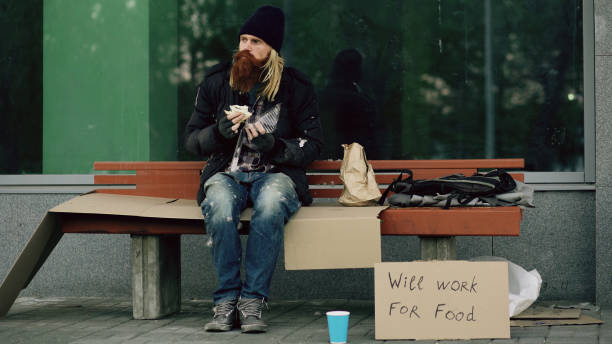 obdachlos und arbeitslos europäischen mann mit pappschild sandwich essen auf bank am stadtstraße wegen einwanderer krise in europa - eating sandwich emotional stress food stock-fotos und bilder