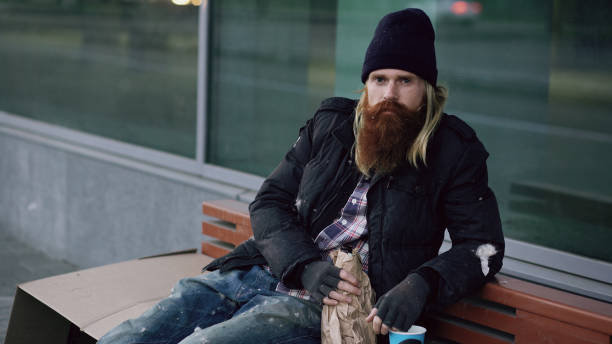 istock drunk homeless sleepy man smiling and looking at the camera while sitting on bench at the sidewalk 892291792