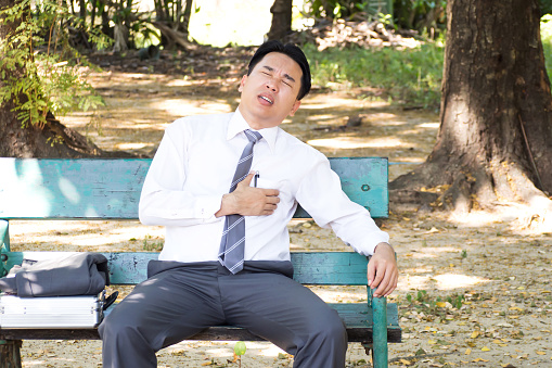 Asian businessman sitting in a park. Chest pain and heart attack. Symptoms of heart disease. Caused by stress and hard work.