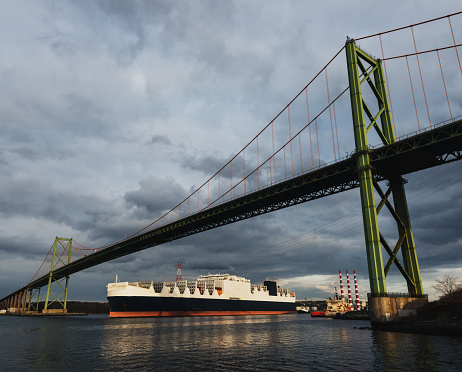 A modern container ship navigates a narrow harbour beneath a suspension bridge.