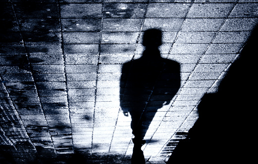 Blurry shadow and silhouette of a man standing in the night on wet city street sidewalk with water reflection