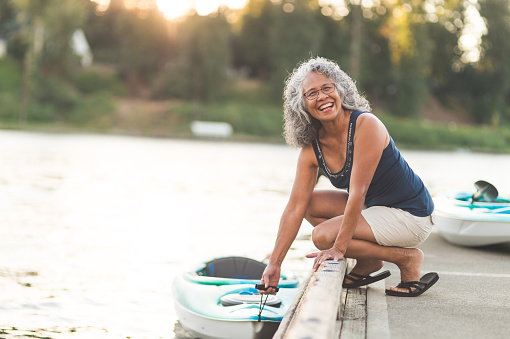 A beautiful older Hawaiian woman prepares to go river kayaking on a summer evening. She is lowering the kayak into the water and looking off-camera and smiling.