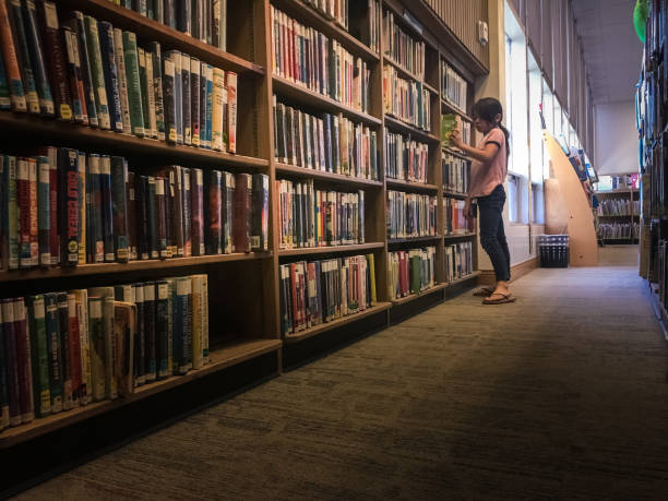 A little girl is standing in a library, searching for a particular book. She's alone in the quiet indoor setting. Taken on an iPhone 6s Plus in Long Beach, CA. The little girl is 7 years old and Eurasian / mixed race.