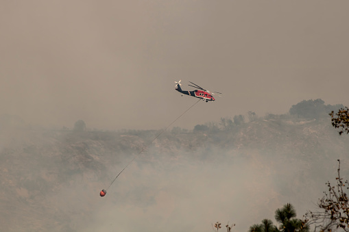 Fire fighting helicopter flies through smoke during Thomas Fire in California with water bucket hanging below.