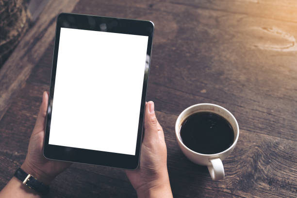 istock Mockup image of business woman's hands holding black tablet pc with blank white screen and coffee cup on vintage wooden table in cafe background 892644170