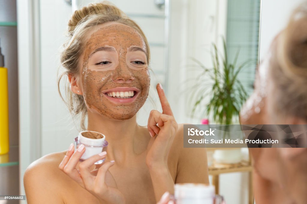 Young beautiful girl applying facial scrub mask on skin. Young beautiful girl applying facial scrub mask on skin. Looking in the mirror in bathroom, Wrapped in a towel, having fun. Facial Mask - Beauty Product Stock Photo